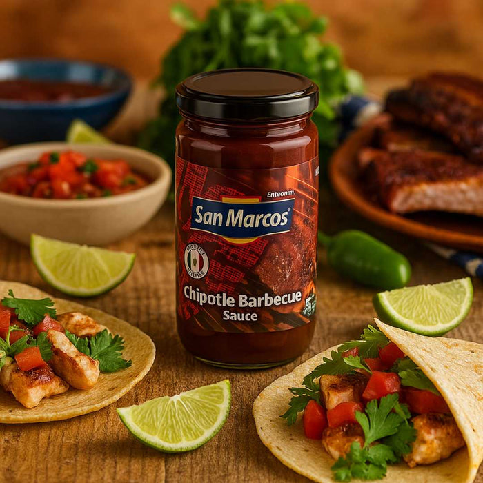 Jar of San Marcos Chipotle Barbecue Sauce on a wooden table with food items like tacos and limes.