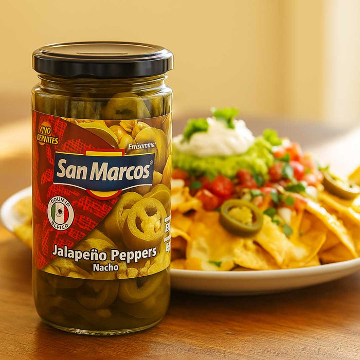 Jar of San Marcos jalapeño peppers next to a plate of nachos with toppings on a wooden surface.