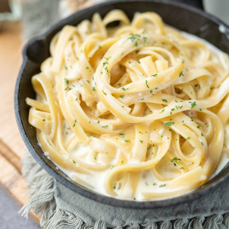 Pasta dish with creamy Knorr sauce and herbs in a black bowl on a wooden surface.